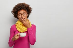 Tired curly woman yawn, has sleepy expression, drinks coffee early in morning, holds white muf of hot beverage, fatigue after work, wears bright stylish clothes, isolated on white wall, empty space